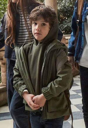 Giacca verde con cappuccio e maniche arricciate sopra una maglia abbinata, indossata con jeans scuri. Tra le figure circostanti ci sono altre due persone in abbigliamento casual.