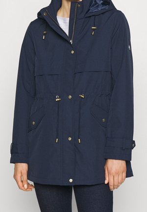 Femme portant une veste à capuche bleu marine avec cordons à la taille et à la capuche, fermetures à boutons-pression et poches à rabat.