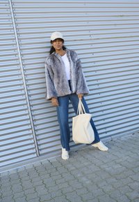 Faux fur jas in grijstinten, witte T-shirt, lichtblauwe spijkerbroek, witte sneakers en beige tote bag. Achtergrond bestaat uit gegolfd metaal.