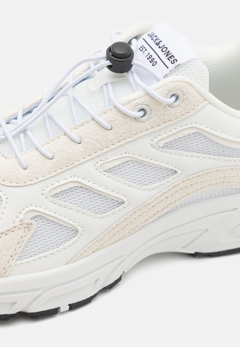 Zapatilla deportiva blanca con parte superior de ante texturizado y malla, que cuenta con un sistema de cordones tipo toggle y una suela de goma negra.