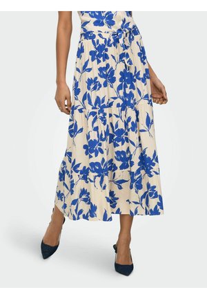 Femme portant une jupe midi plissée couleur crème avec un imprimé floral bleu vif, associée à des escarpins pointus bleu foncé.