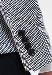 Närobild av en ärm på en blårandig kavaj med fyra svarta knappar över en vit skjortärm.