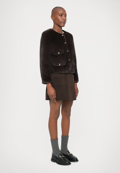 Brun syntetisk pelsjakke med to frontlommer og gullfargede knapper, i kombinasjon med en brun semsket skinnskjørt og svarte loafers med grå sokker.