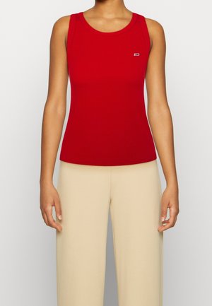 Débardeur rouge côtelé avec un col rond et des larges bretelles, orné d'un petit logo sur la poitrine. Associé à un pantalon beige à jambes larges.