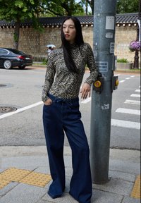 Maglietta a maniche lunghe con motivo a strisce zebra nera e beige, abbinata a jeans in denim blu scuro a vita alta; il modello si appoggia a un palo della luce.