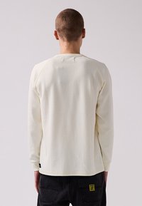 Long-sleeve shirt in light cream, featuring a textured waffle pattern, raglan sleeves, and a small logo tag near the hem.