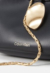 Černá kožená kabelka se zlatým kovovým akcentem, vybavená texturovaným řetízkovým popruhem a vyrytým logem "Calvin Klein".