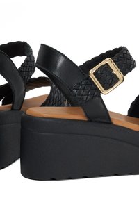 Zwarte platform sandalen met geweven banden en een gouden sluiting. Kenmerken zijn een gestructureerde zwarte zool en een gladde, tan kleurige voetbed.