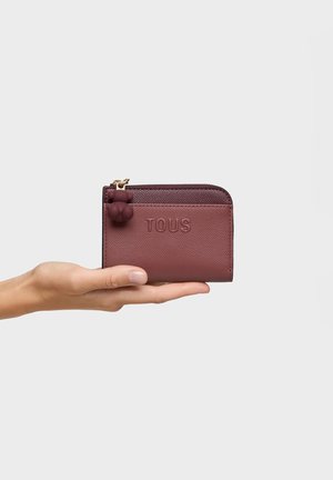 Hand holding a small maroon wallet with embossed "TOUS" text and a bear-shaped zipper pull on a plain light background.