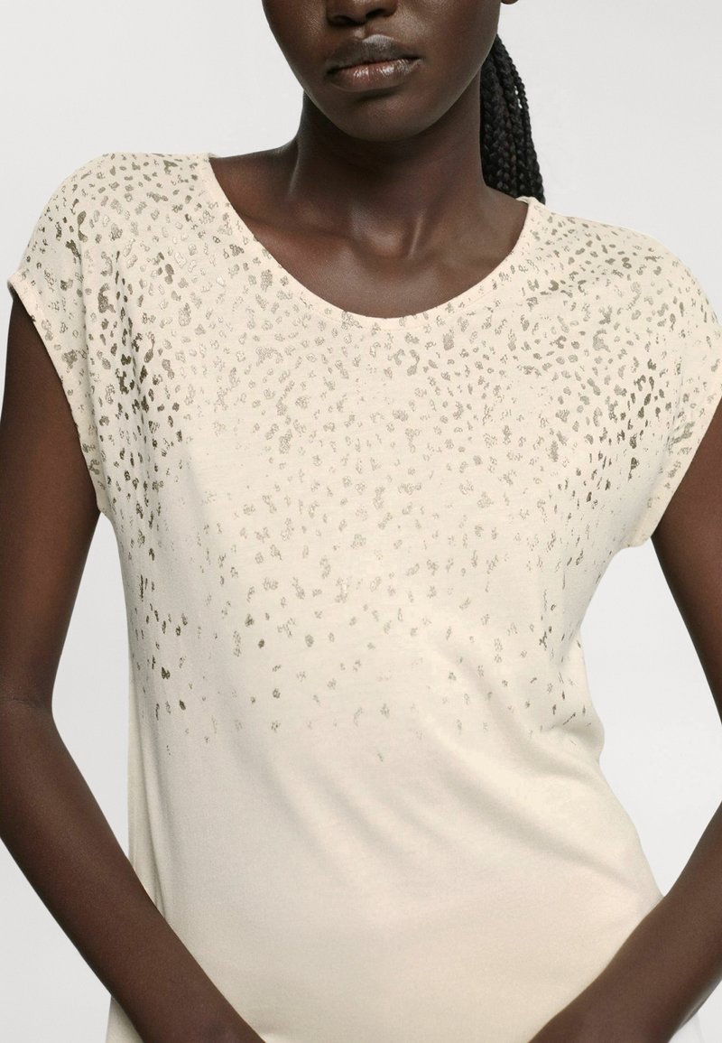 Krémový top s krátkým rukávem a kulatým výstřihem, ozdobený texturovanými metalickými tečkami a hladkým materiálem, s volným střihem.