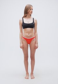 Νεαρή γυναίκα όρθια, ξυπόλυτη, φορώντας μαύρο αθλητικό σουτιέν και κόκκινο κάτω μέρος μπικίνι, κοιτάζει προς τα αριστερά της με φόντο ένα λευκό, απλό φόντο.