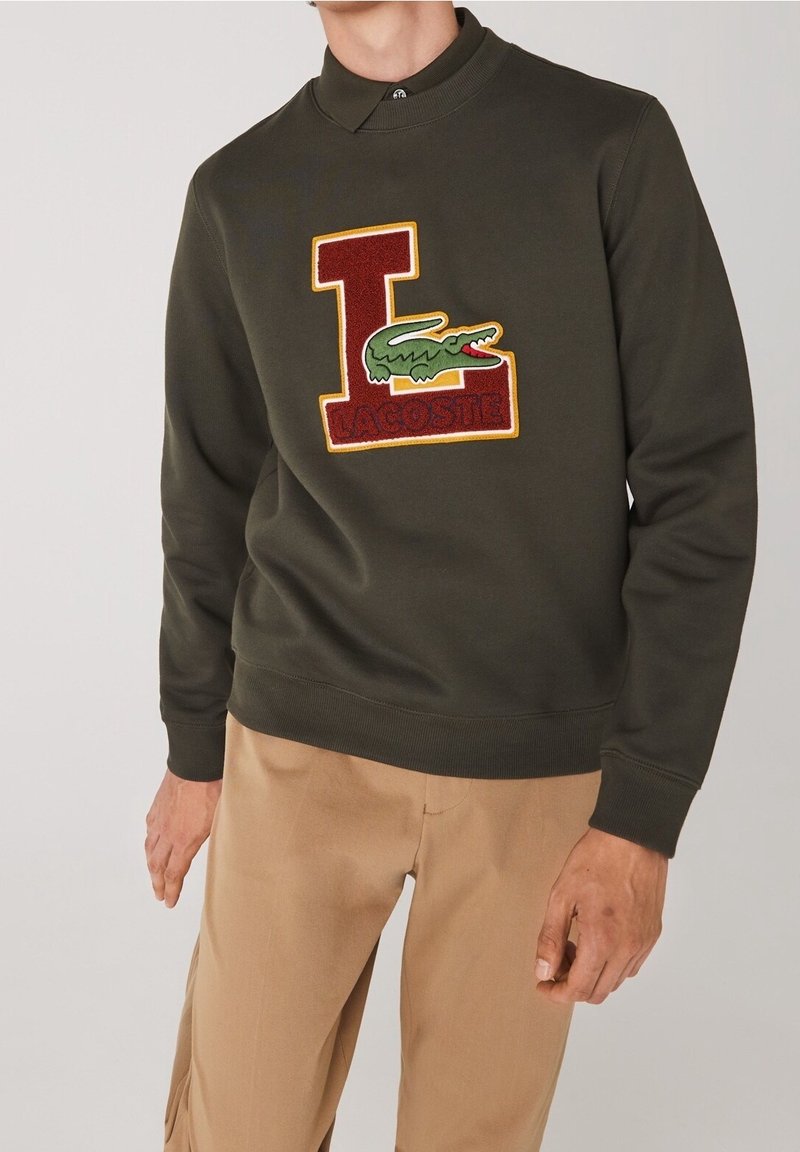 Homme portant un sweat-shirt vert foncé avec un grand « L » rouge et jaune et un logo crocodile vert, associé à un pantalon beige.
