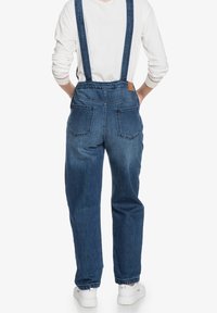 Jeansoverall i mörkblått med två justerbara axelremmar, bakfickor och en brun läderetikett. Vit långärmad skjorta och sneakers.