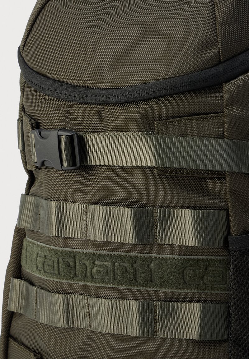 Zaino in tessuto verde con superficie strutturata, spallacci regolabili, fibbia e una vistosa toppa con il logo.