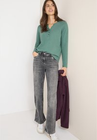 Vihreä pitkähihainen henley-paita, jossa on kolme nappia, yhdistettynä harmaisiin leveälahkeisiin farkkuihin. Malli pitää kädessään purplea takkia ja hänellä on päällään valkoiset tennarit.