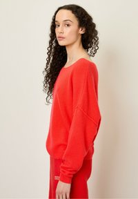 Lyse rød oversized sweater med en blød tekstur, bred halsudskæring og sænkede skuldre. Matchet med røde bukser i samme farve.