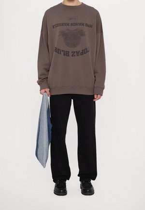 Henkilö, joka pukeutunut ylisuurikokoiseen ruskeaan collegepaitaan, jossa musta teksti, mustat housut, mustat kengät, ja pitää toisessa kädessään sinivalkoista kangasta.