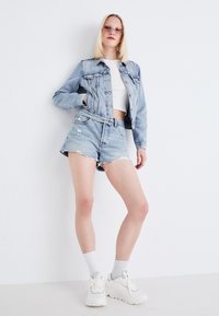 Ljusblå denimjacka, vit kortärmad topp, mörka denimshorts, ihop med vita sneakers och grå strumpor, mot en ren vit bakgrund.