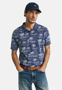 Marineblauwe poloshirt met een wit patroon van palmbomen en golven, korte mouwen, kraag, gecombineerd met vervaagde denim jeans en een bruine riem.