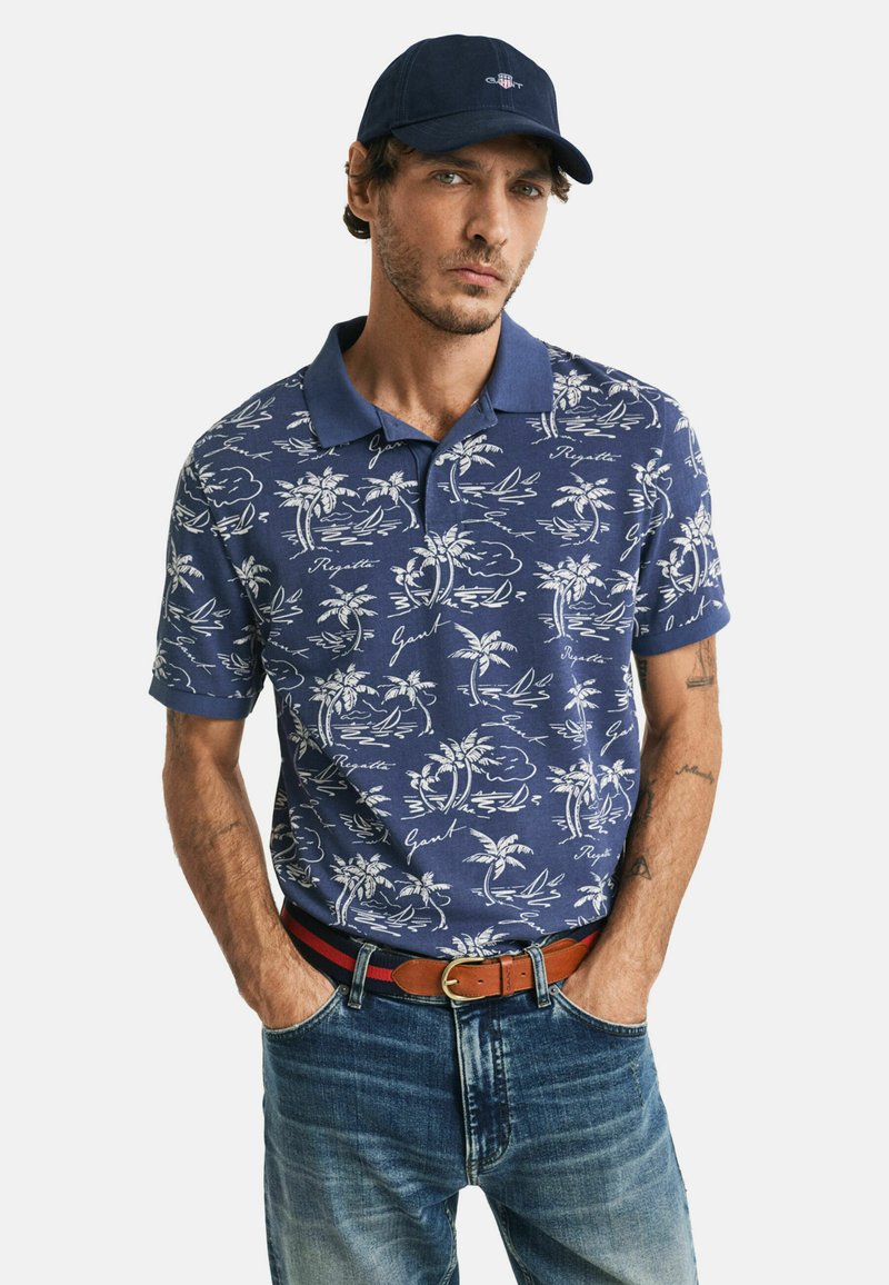 Marineblauwe poloshirt met een wit patroon van palmbomen en golven, korte mouwen, kraag, gecombineerd met vervaagde denim jeans en een bruine riem.