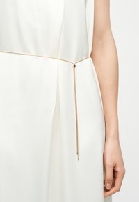 Λεπτή χρυσή ζώνη-αλυσίδα με μικρή μαύρη πέτρα ως διακόσμηση, φορεμένη πάνω από λευκό αμάνικο φόρεμα, εμφανίζεται στη δεξιά πλευρά του κορμού ενός ατόμου.