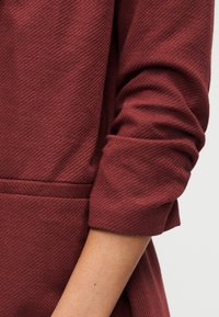 Μανίκι γυρισμένο πάνω σε υφασμάτινο, μακρυμάνικο ένδυμα σκούρου κόκκινου χρώματος, που δείχνει λεπτομέρειες του υφάσματος και μέρος του βραχίονα.