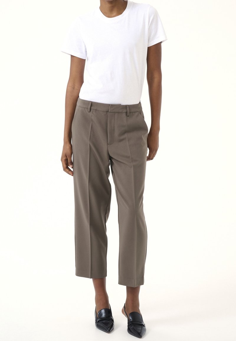 Personne portant un t-shirt blanc ajusté à manches courtes, un pantalon taupe court taille haute et des chaussures noires à bout pointu avec bride arrière, debout.