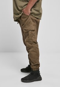 Khaki cargo kalhoty s elastickými manžetami, několika kapsami a volným střihem, spárované s černými sportovními botami. Matná textura, odolný materiál.