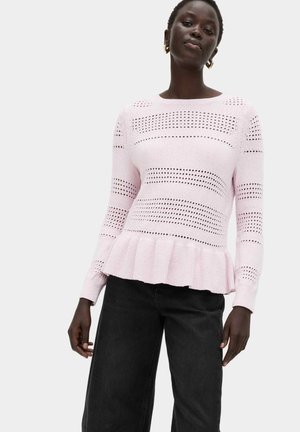 Vrouw draagt een lichtroze gebreide trui met geperforeerde horizontale strepen en een gerimpelde zoom, gecombineerd met een zwarte broek, staand tegen een witte achtergrond.