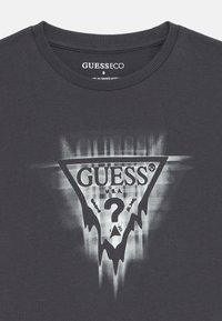 Pilkas medvilninis marškinėliai su apvalia iškirpte, turintis trikampį grafikos dizainą, kuriame yra žodis "GUESS", klausimo ženklas ir nusidėvėję akcentai.