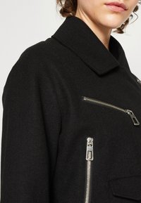 Manteau noir en mélange de laine avec un grand col, poche poitrine zippée et poches latérales avec des accents en métal argenté.