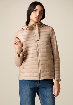 Donna che regola il laccio di una giacca trapuntata beige, indossa jeans blu, in piedi davanti a uno sfondo chiaro e uniforme.