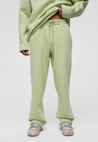 Lichtgroene sweatpants met een elastische tailleband, verstelbare koord, en zijzakken; gecombineerd met beige sneakers. Zachte stoftextuur.