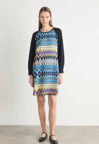 Vestido colorido con patrones geométricos en azul, naranja y negro. Presenta mangas largas negras y un escote redondo, confeccionado con tela estampada.