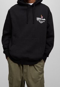 Czarna bluza z kapturem z przednią kieszenią, małe kolorowe logo i napis po lewej górnej stronie, w połączeniu z oliwkowozielonymi spodniami typu cargo.