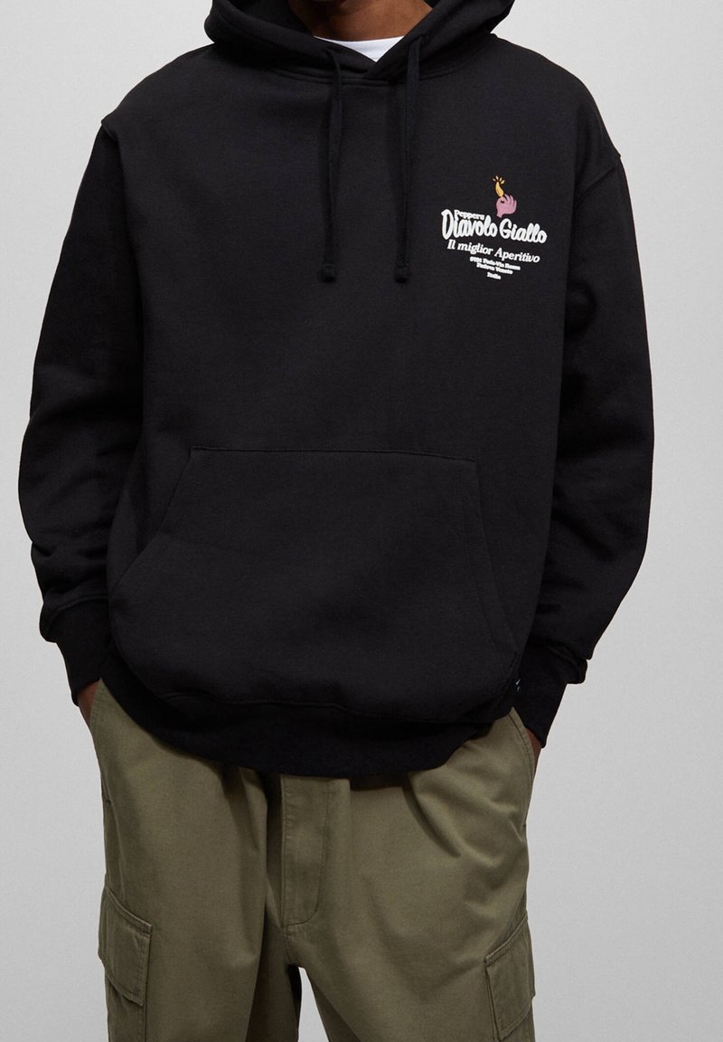 Czarna bluza z kapturem z przednią kieszenią, małe kolorowe logo i napis po lewej górnej stronie, w połączeniu z oliwkowozielonymi spodniami typu cargo.