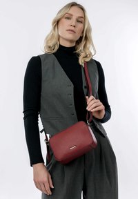 Raudona odinė kryžminė šoninė rankinė su tekstūruota paviršiumi, stačiakampio formos ir auksiniu logotipo akcentu. Reguliuojama dirželis, elegantiškas dizainas.