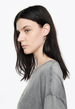 Camiseta gris de gran tamaño con una textura deslavada; presenta un cuello redondo y mangas cortas. Pendientes de aro dorados con un diseño de hoja.