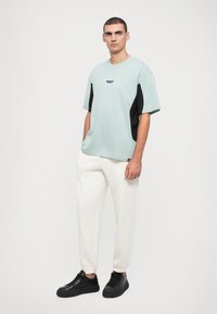 Μαint πράσινη oversized μπλούζα με μαύρες πλαϊνές ρίγες και μικρή λεπτομέρεια λογότυπου, συνδυασμένη με λευκά tapered παντελόνια και μαύρα αθλητικά παπούτσια.