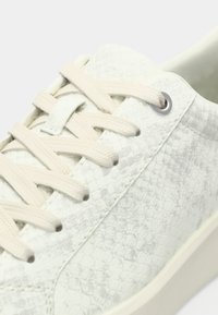 Gros plan sur une chaussure de sport blanche avec un motif texturé façon peau de serpent et des lacets beiges passés dans des œillets en métal.