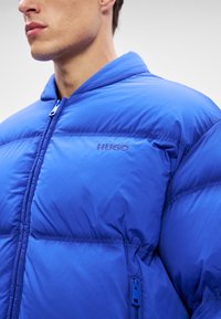 Mėlynas pūkinis švarkas, pagamintas iš sintetinių medžiagų, su užtrauktuku priekyje, stovinčiu apykakle ir horizontaliais pūkų raštais. Logotipas ant krūtinės.