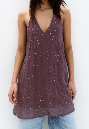 Femme portant une robe violette sans manches à petits pois blancs sur un jean bleu clair, debout devant un fond blanc uni.