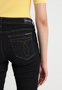 Vaqueros de mezclilla negros con un diseño ajustado, que presentan costuras naranjas en contraste y una etiqueta de logo en la cintura y en el bolsillo trasero.