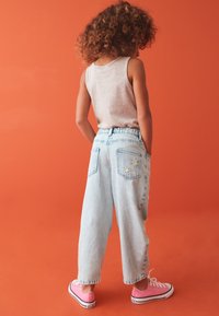 Pantalones de mezclilla azul claro con cintura elástica, dos bolsillos traseros con bordados florales, combinados con zapatillas rosas, sobre un fondo naranja.