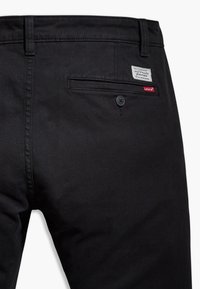 I pantaloni in denim neri presentano una tasca posteriore con bottone e una piccola etichetta. Il tessuto ha una consistenza liscia con una leggera lucentezza.