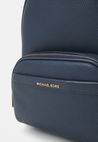 Zaino in pelle texturizzata blu navy con zip dorata e accento del logo; presenta una forma arrotondata e una tasca frontale con chiusura a zip.