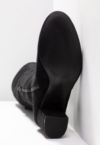 Botte mi-mollet en daim noir avec talon bloc, fermeture éclair sur le côté et semelle en caoutchouc texturée avec motif ondulé vue de dessous.