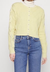 Cardigan giallo con texture e motivo a diamante, con bottoni perla e maniche decorate, indossato su un dolcevita bianco e jeans blu.