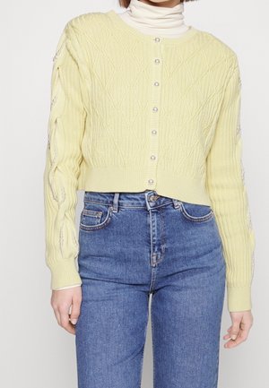 Gul tekstureret cardigan med diamantmønster, prydet med perleknapper og udsmykkede ærmer, båret over en hvid turtleneck og blå jeans.