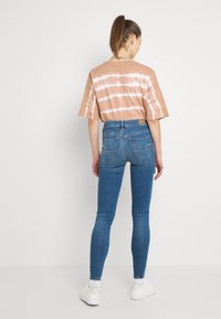 Brun tie-dye kortärmad skjorta i kombination med blå jeans i skinny-stil och vita sneakers. Jeanserna har bakfickor och en läderlapp.